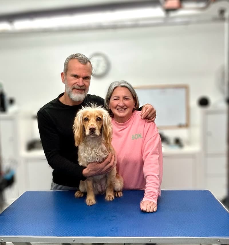 The two groomers standing behind a freshly groomed cocker spaniel on a blue grooming table in their salon